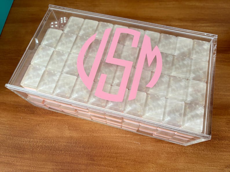 Op de afbeelding: Een doorzichtige acrylbox met een roze monogram op het deksel. De doos is gevuld met kleine, vierkante, parelmoerachtige objecten. De doos staat op een houten oppervlak.