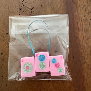 May include: Three pink Mahjong tile charms, numbered 1, 2, and 3, are displayed in a clear plastic bag. Each tile features a different design in shades of blue and green. A blue ribbon is attached to the top of the tiles.