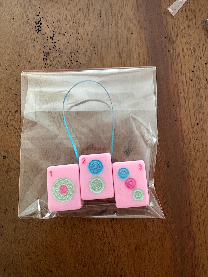 May include: Three pink Mahjong tile charms, numbered 1, 2, and 3, are displayed in a clear plastic bag. Each tile features a different design in shades of blue and green. A blue ribbon is attached to the top of the tiles.
