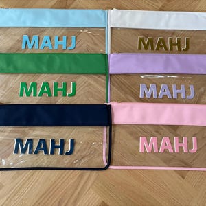 May include: Clear zippered pouches with colored tops in light blue, white, green, lavender, navy, and pink. Each pouch has the letters "MAHJ" in a coordinating color. The pouches are arranged on a light wood surface.