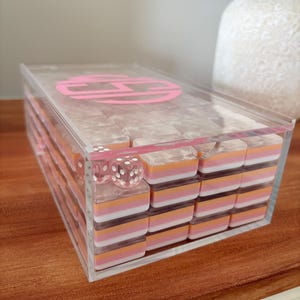 Op de afbeelding: Een doorzichtige acryldomino set met roze, oranje en witte dominostenen. De set bevat twee dobbelstenen en wordt bewaard in een doorzichtige doos met een roze monogram op het deksel. De dominostenen zijn netjes in de doos gestapeld.