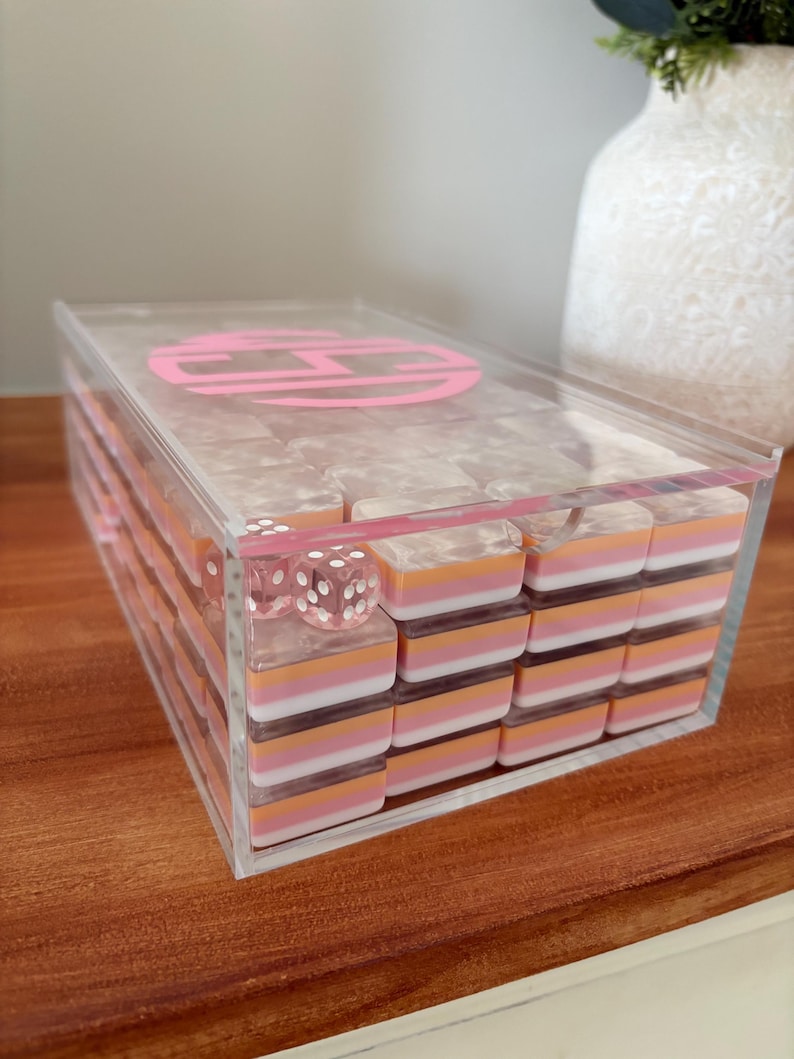 Op de afbeelding: Een doorzichtige acryldomino set met roze, oranje en witte dominostenen. De set bevat twee dobbelstenen en wordt bewaard in een doorzichtige doos met een roze monogram op het deksel. De dominostenen zijn netjes in de doos gestapeld.