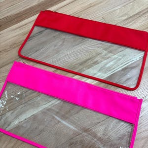 May include: Two rectangular zipper pouches, one in bright red and one in neon pink. Each pouch features a clear transparent plastic lower section and a solid colored fabric upper section with a matching zipper closure along the top edge.