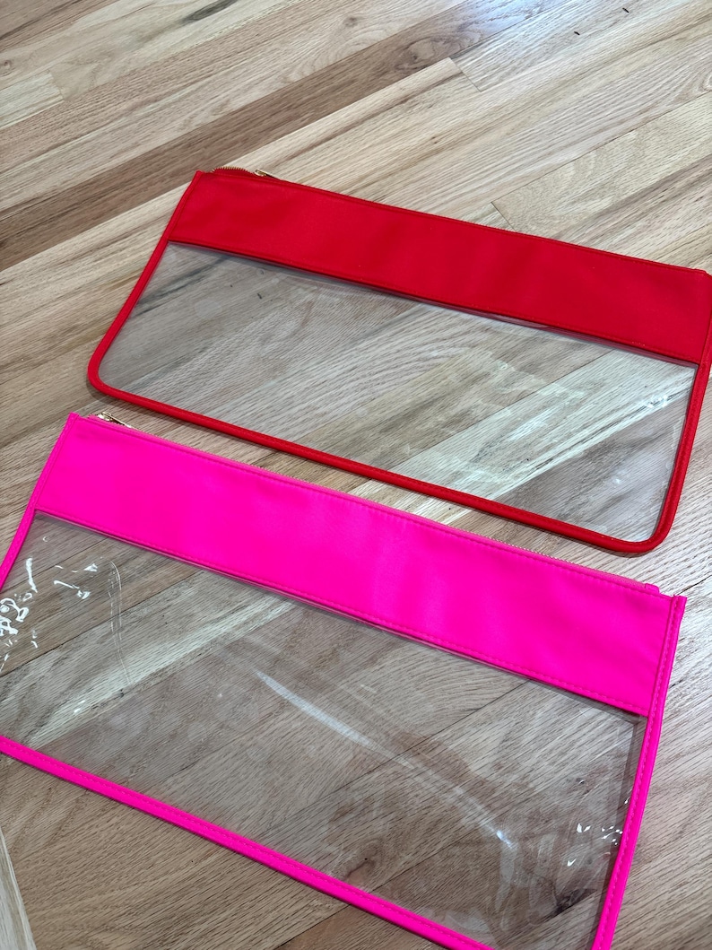 May include: Two rectangular zipper pouches, one in bright red and one in neon pink. Each pouch features a clear transparent plastic lower section and a solid colored fabric upper section with a matching zipper closure along the top edge.