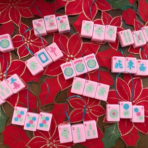 May include: Pink Mahjong tile ornaments with various symbols and numbers, strung with blue ribbon. The ornaments are arranged on a red poinsettia-themed background, creating a festive holiday display.