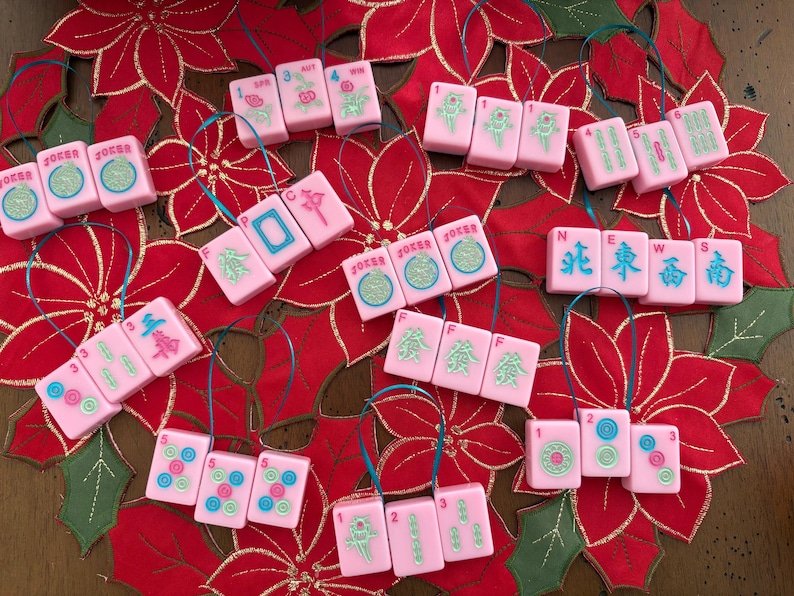 May include: Pink Mahjong tile ornaments with various symbols and numbers, strung with blue ribbon. The ornaments are arranged on a red poinsettia-themed background, creating a festive holiday display.
