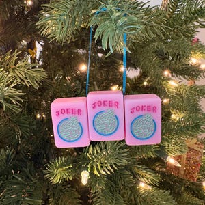 May include: Three pink Christmas ornaments shaped like dice, each with the word "JOKER" and a blue and green design. The ornaments hang from a blue string against a backdrop of a decorated Christmas tree.
