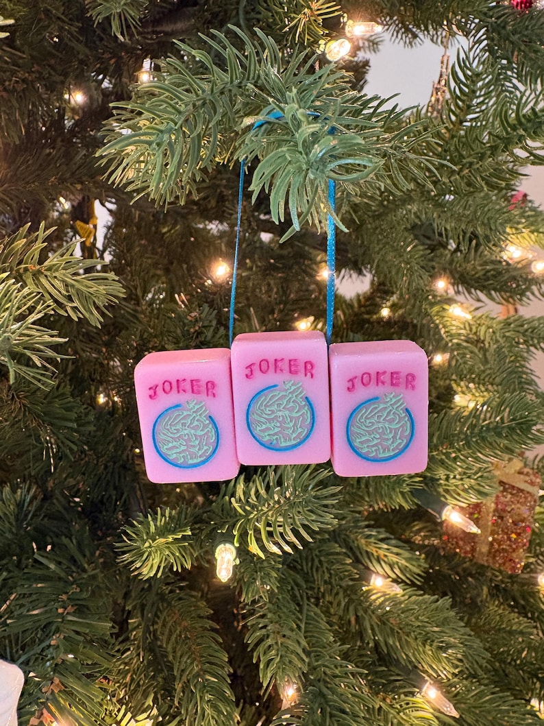 May include: Three pink Christmas ornaments shaped like dice, each with the word "JOKER" and a blue and green design. The ornaments hang from a blue string against a backdrop of a decorated Christmas tree.