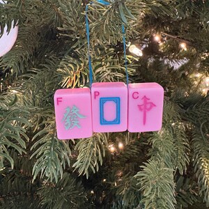 May include: A pink Mahjong tile Christmas ornament. The ornament is made of three pink tiles with Chinese characters and symbols in blue and green. The ornament hangs from a blue ribbon against a backdrop of a Christmas tree.
