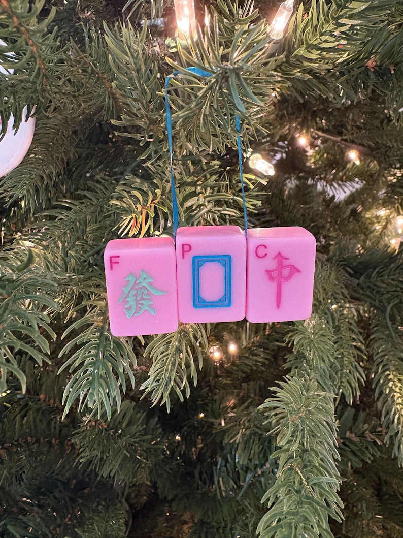 May include: A pink Mahjong tile Christmas ornament. The ornament is made of three pink tiles with Chinese characters and symbols in blue and green. The ornament hangs from a blue ribbon against a backdrop of a Christmas tree.