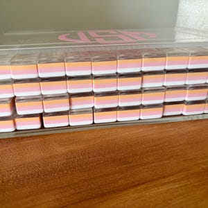 Op de afbeelding: Een doorzichtige acrylbox gevuld met rijen kleine, rechthoekige gummen. Elke gum heeft een wit, roze en oranje kleurenschema. De doos heeft een roze monogram op het deksel en staat op een houten oppervlak.