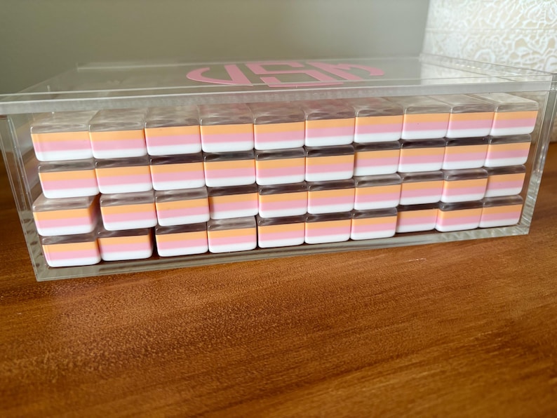 Op de afbeelding: Een doorzichtige acrylbox gevuld met rijen kleine, rechthoekige gummen. Elke gum heeft een wit, roze en oranje kleurenschema. De doos heeft een roze monogram op het deksel en staat op een houten oppervlak.