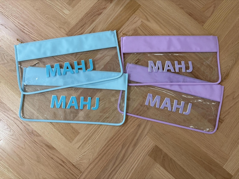 May include: Four rectangular zipper pouches made of clear plastic with colored borders and matching lettering. Two pouches feature light blue trim and the text "MAHJ" in light blue, while two feature lavender trim and the text "MAHJ" in lavender.