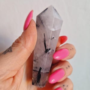 May include: A clear quartz crystal point with black tourmaline inclusions. The crystal is held in a hand with bright pink fingernails. The crystal is a light, translucent colour with dark, linear inclusions.