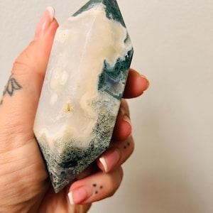 May include: A double-terminated moss agate crystal point featuring a smooth, polished surface with a blend of milky white, translucent areas and deep green, moss-like dendritic inclusions throughout the stone's elongated, hexagonal structure.
