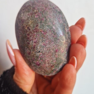 May include: A rounded, oval stone with a mottled appearance. The stone's surface is a blend of grey, green, and pink tones, with small, reddish-pink inclusions. The stone is held in a hand, highlighting its polished texture.