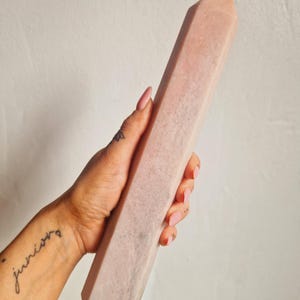 May include: A light pink crystal obelisk, held in a hand. The obelisk is rectangular with a pointed top. The crystal has a textured surface and a soft, muted pink color. The word "juniors" is tattooed on the wrist.