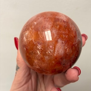 Könnte beinhalten: Eine polierte, kugelförmige Edelstein-Kugel in Orange- und Rottönen. Die Oberfläche reflektiert das Licht und zeigt interne Muster und Einschlüsse. Diese dekorative Kugel ist ein einzigartiges Wohnaccessoire. Durchmesser ca. 7 cm.