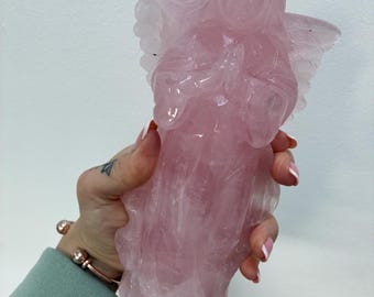 May include: A carved rose quartz angel figurine with outstretched wings. The angel is a soft pink color and has detailed facial features and flowing robes. The figurine is held in a hand against a white background.