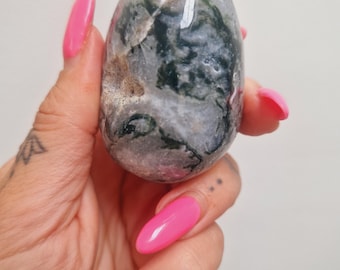 May include: A polished, oval-shaped gemstone with a mix of gray, green, and brown hues. The stone has a smooth surface and a natural, organic appearance. The gemstone is held in a hand with bright pink fingernails.