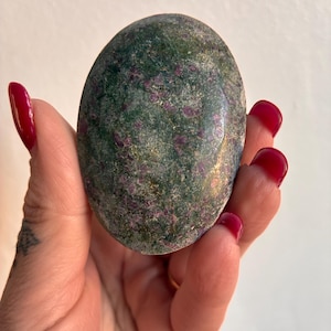 May include: A smooth, oval-shaped stone with a mottled green and red pattern. The stone is held in a hand with red nail polish. The stone's surface has a slightly polished appearance, with the red inclusions adding visual interest.