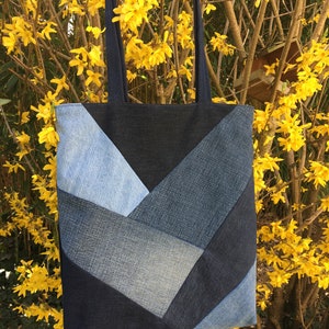 Peut inclure: Un sac fourre-tout en denim avec un motif en chevrons dans les tons de bleu. Le sac a deux anses bleu foncé et est suspendu devant un fond de fleurs jaunes et de branches vertes. Le sac est fait de denim recyclé.