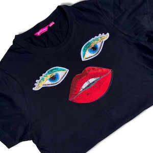 Op de afbeelding: Zwart T-shirt met pailletten ogen en lippen applicaties. De ogen hebben blauwe irissen, gouden oogleden en groene en witte accenten. De lippen zijn rood met zilveren pailletten. Het shirt heeft een ronde hals en een label "JPS wear".