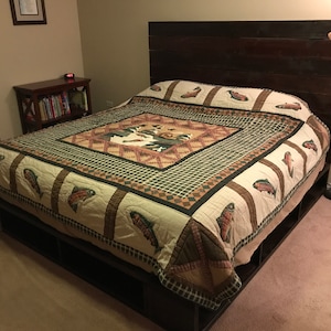 May include: A wooden bed frame with a dark brown headboard and a storage platform. The bed is made with a green and brown patterned quilt with a fish design.