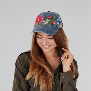 May include: A woman wearing a blue denim baseball cap with colorful embroidered flowers. The cap has a curved brim and a slightly distressed look. She is wearing a dark green shirt with a button-up collar.