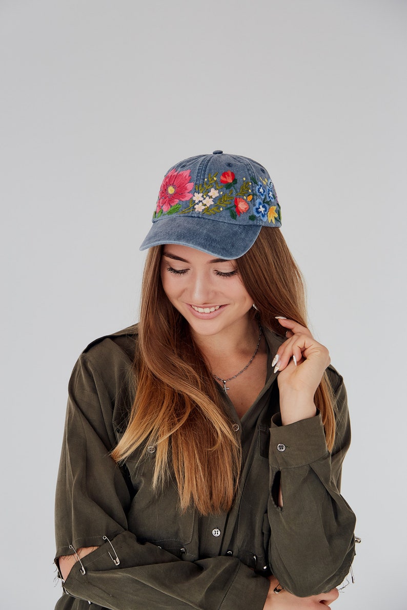 May include: A woman wearing a blue denim baseball cap with colorful embroidered flowers. The cap has a curved brim and a slightly distressed look. She is wearing a dark green shirt with a button-up collar.