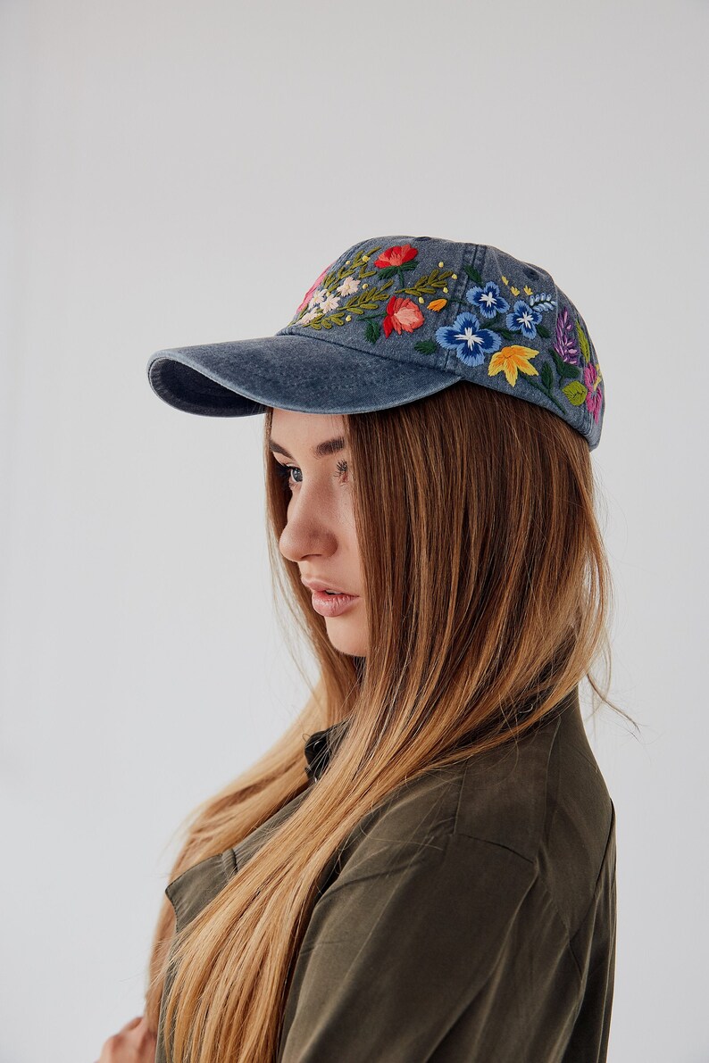 May include: A woman with long blonde hair is wearing a blue denim baseball cap with colorful embroidered flowers. The cap has a curved brim and a white adjustable strap.