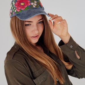 May include: A woman with long brown hair is wearing a blue denim baseball cap with colorful embroidered flowers. She is wearing a dark green shirt with buttons and a pocket.