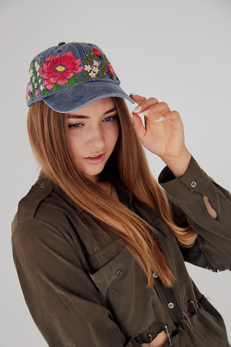 May include: A woman with long brown hair is wearing a blue denim baseball cap with colorful embroidered flowers. She is wearing a dark green shirt with buttons and a pocket.