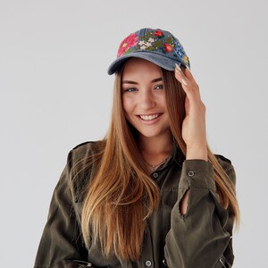 May include: A woman with long brown hair is wearing a green shirt with a blue baseball cap with embroidered flowers. She is smiling and looking at the camera.