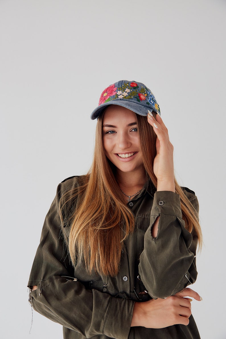 May include: A woman with long brown hair is wearing a green shirt with a blue baseball cap with embroidered flowers. She is smiling and looking at the camera.