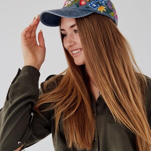 May include: A woman wearing a blue denim baseball cap with colorful embroidered flowers. The cap has a curved brim and a mesh back. She is wearing a dark green shirt with safety pins holding the fabric together.