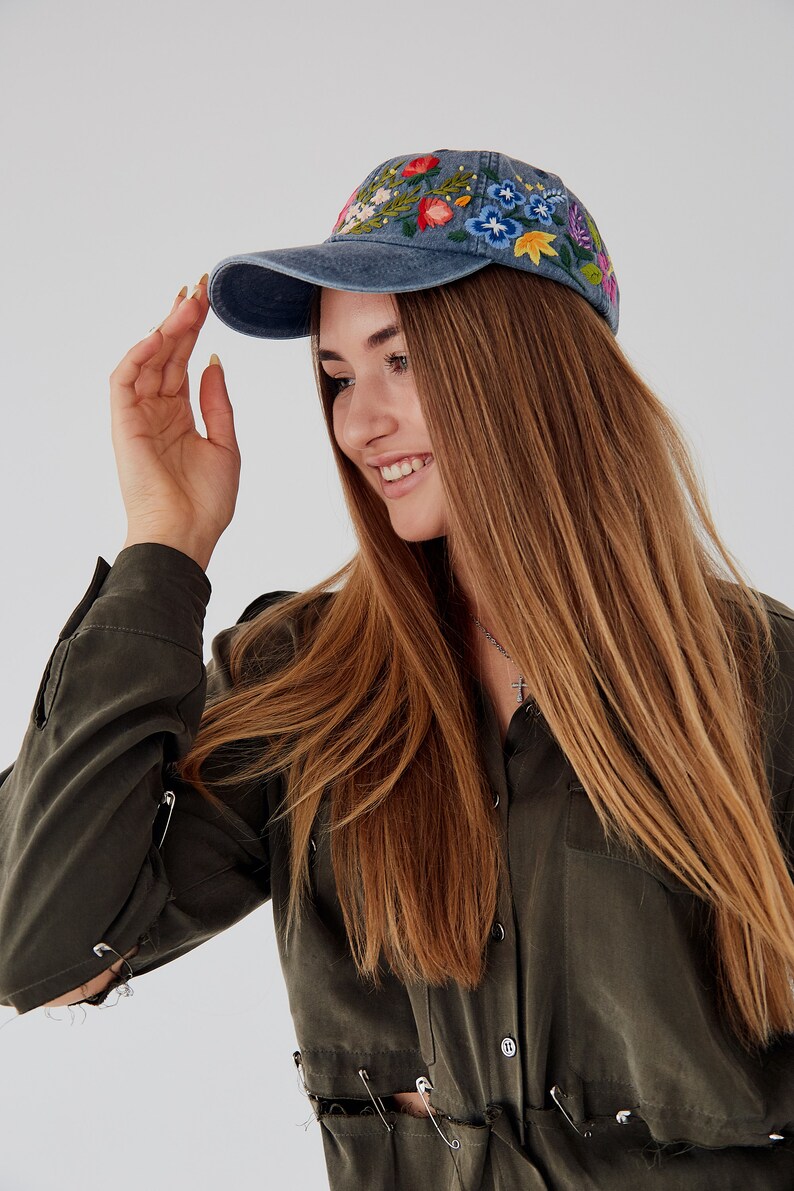 May include: A woman wearing a blue denim baseball cap with colorful embroidered flowers. The cap has a curved brim and a mesh back. She is wearing a dark green shirt with safety pins holding the fabric together.