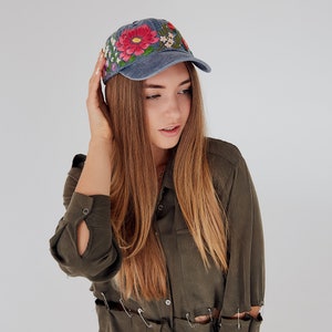 May include: A woman wearing a blue denim baseball cap with embroidered flowers and a green button-down shirt with safety pins holding the fabric together.
