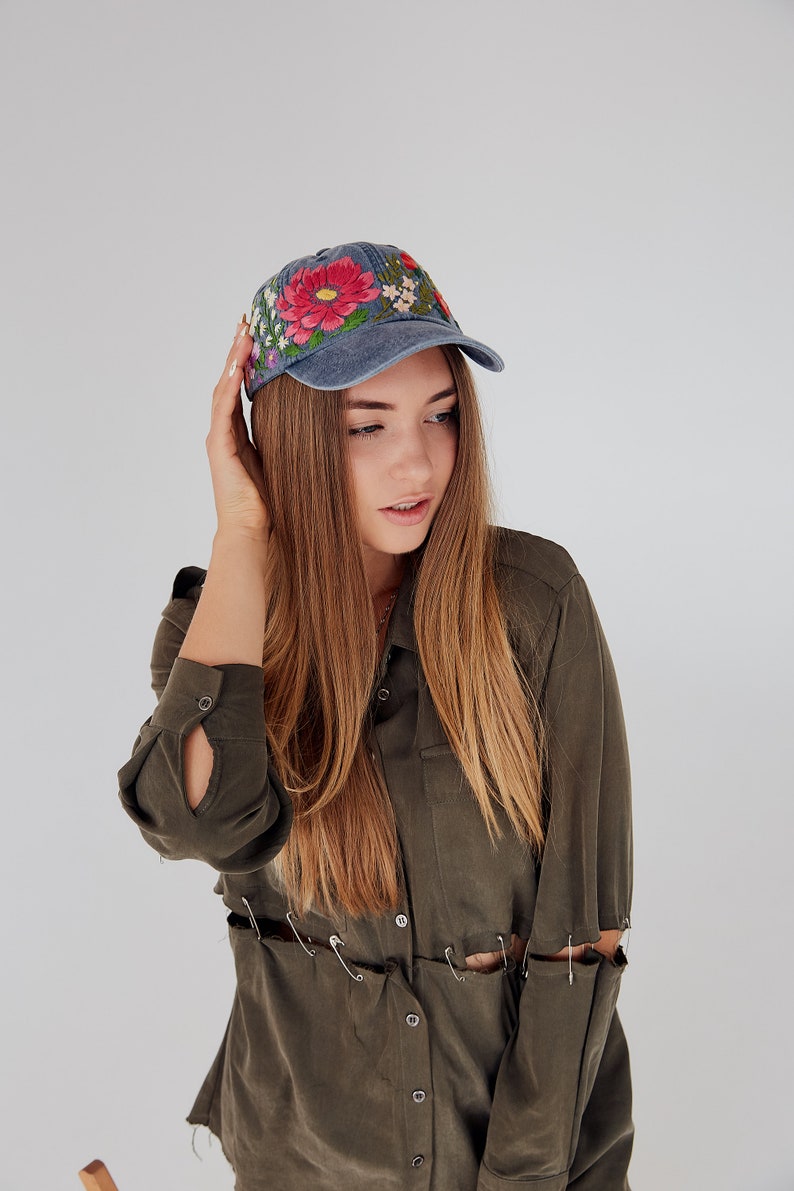 May include: A woman wearing a blue denim baseball cap with embroidered flowers and a green button-down shirt with safety pins holding the fabric together.