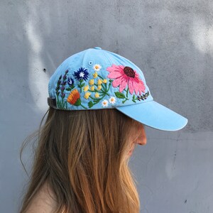 May include: A light blue baseball cap with embroidered flowers in shades of pink, orange, yellow, blue, and green.