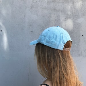May include: A light blue baseball cap with a brown adjustable strap. The cap is being worn by a person with long blonde hair.