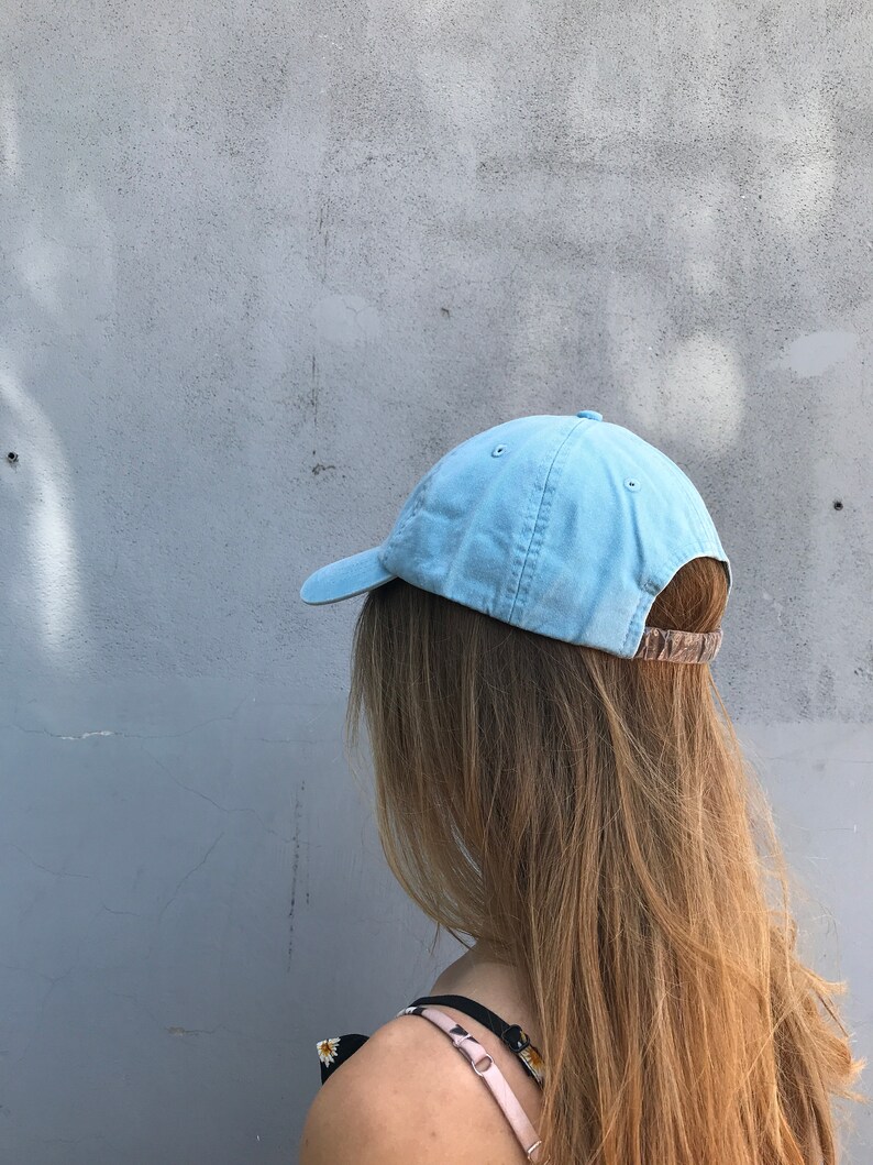 May include: A light blue baseball cap with a brown adjustable strap. The cap is being worn by a person with long blonde hair.