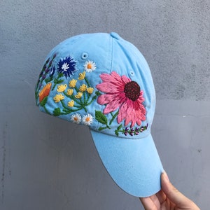 May include: A light blue baseball cap with a colourful embroidered floral design. The flowers are pink, yellow, orange, white, blue, and green.