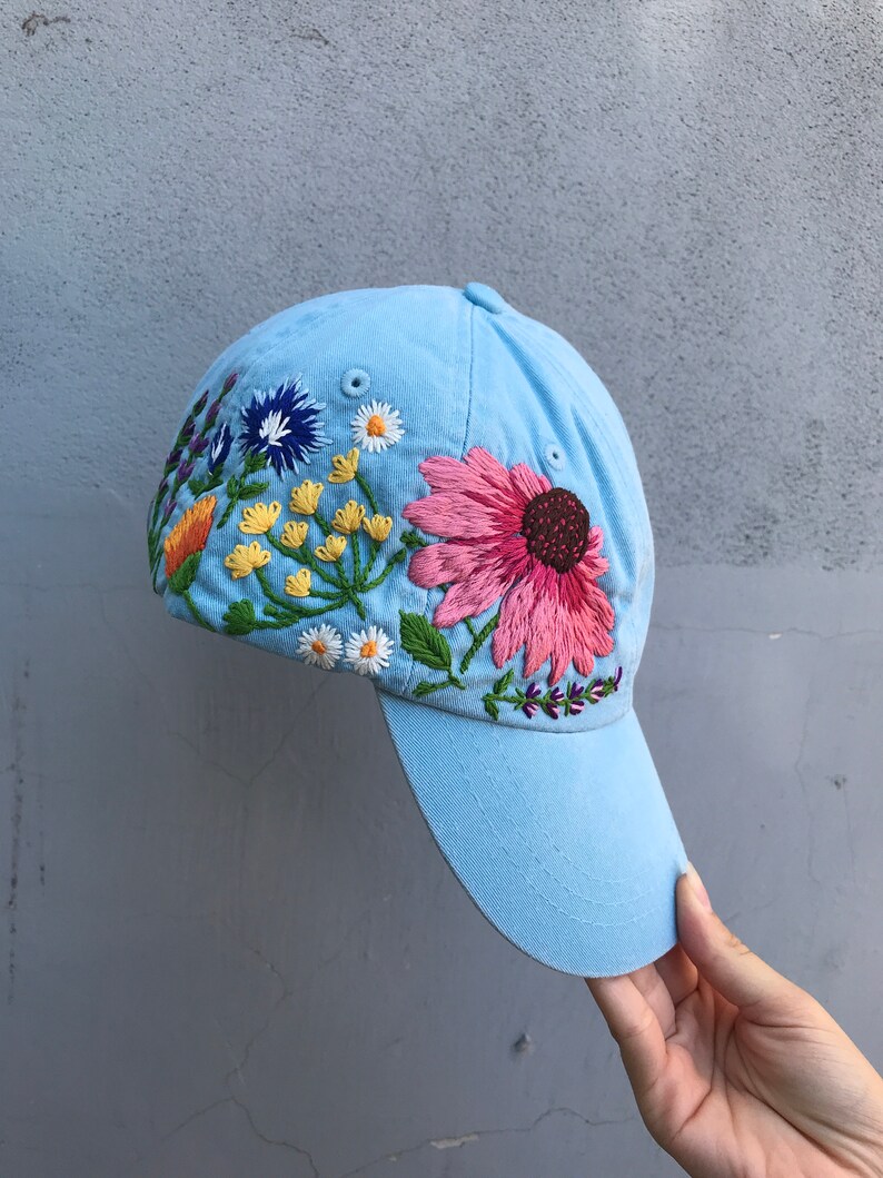 May include: A light blue baseball cap with a colourful embroidered floral design. The flowers are pink, yellow, orange, white, blue, and green.