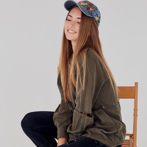 May include: A young woman wearing a blue denim cap with floral embroidery, a green shirt, and blue jeans sits on a wooden chair.