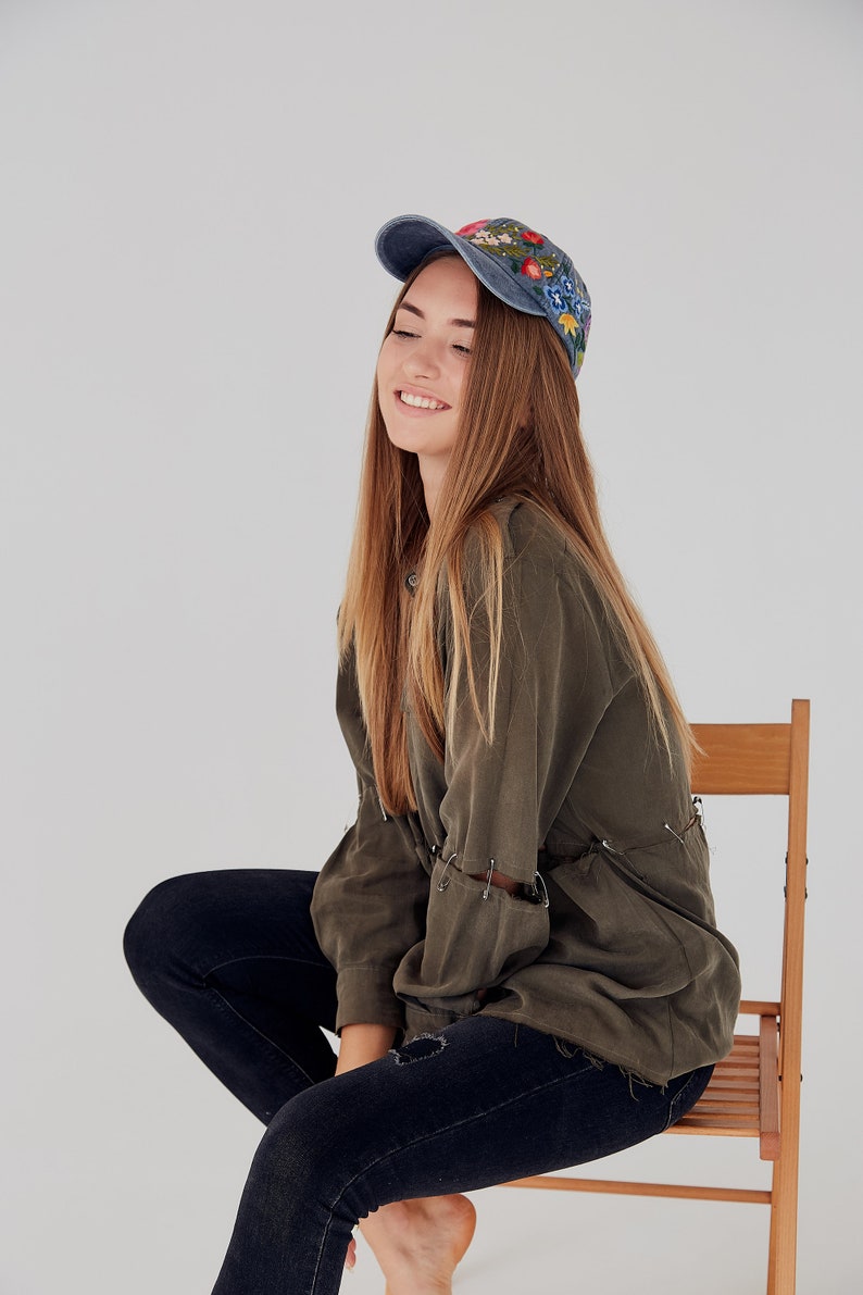 May include: A young woman wearing a blue denim cap with floral embroidery, a green shirt, and blue jeans sits on a wooden chair.