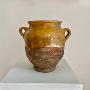 May include: A weathered ceramic pot with two handles. The top section is a bright yellow, fading to a lighter band, and then to a deep brown base. The pot is displayed on a white surface.