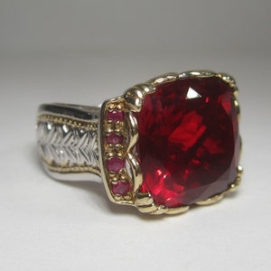 May include: A silver and gold ring with a large red gemstone and smaller red gemstones around the setting.