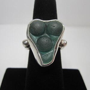 May include: A silver ring featuring a triangular-shaped setting with three dark green, circular, textured stone inlays arranged in a cluster. The metal band has a smooth, polished finish with small spherical accents on each side of the setting.