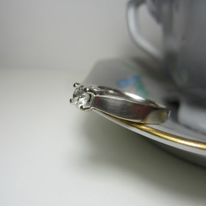 May include: A silver ring with a single round diamond set in the center. The ring is resting on a white saucer with a gold rim.